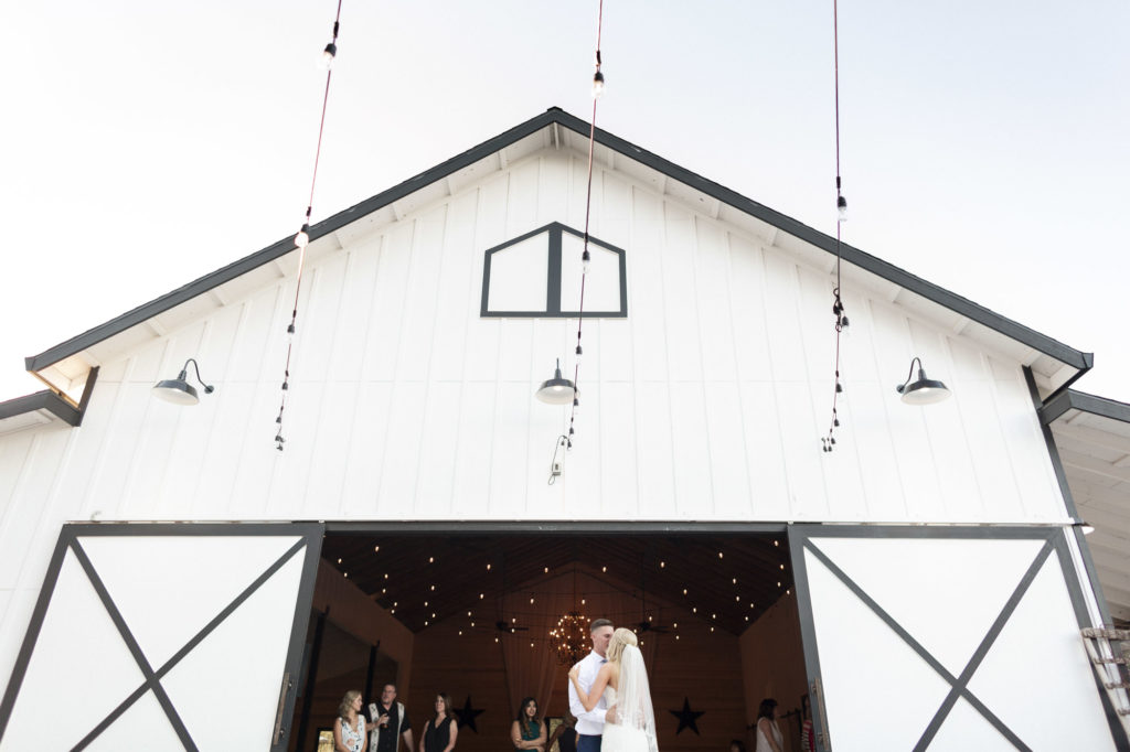 bride and groom dancing in front of white barn with string lights